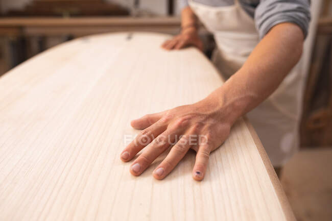 Sezione intermedia del produttore di tavole da surf maschile che lavora nel suo studio, costruisce una tavola da surf, la ispeziona e si prepara alla lucidatura.. — Foto stock