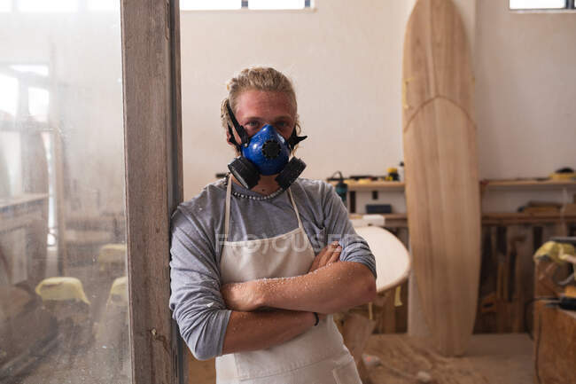 Retrato de um homem caucasiano fabricante de pranchas de surf com longos cabelos loiros, usando uma máscara facial, de pé em seu estúdio com os braços cruzados e olhando para a câmera . — Fotografia de Stock