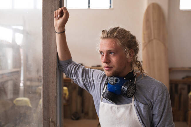 Kaukasischer Surfbrettmacher mit langen blonden Haaren, Gesichtsmaske, steht in seinem Atelier, lehnt an einem Türrahmen, während er während seiner Arbeit eine Pause einlegt. — Stockfoto