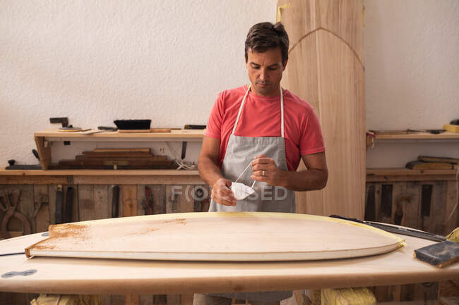 Creatore caucasico di tavole da surf maschile che lavora nel suo studio, indossa un grembiule protettivo, indossa una maschera per il viso e si prepara a lucidare una tavola da surf. — Foto stock