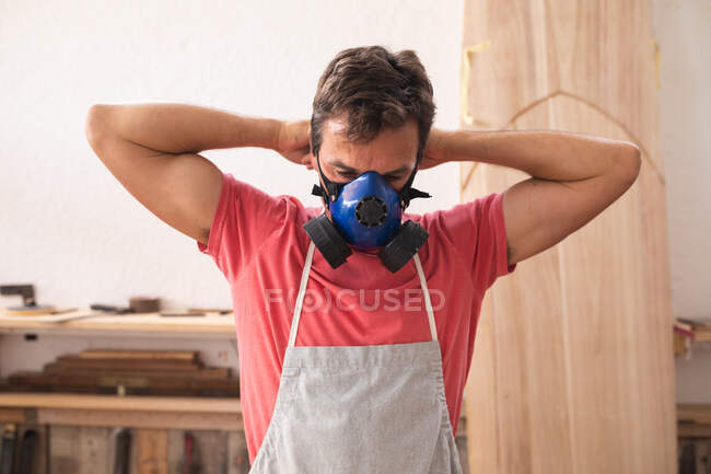 Homem caucasiano fabricante de pranchas de surf trabalhando em seu estúdio, vestindo um avental protetor, colocando uma máscara facial respirando se preparando para polir uma prancha de surf . — Fotografia de Stock