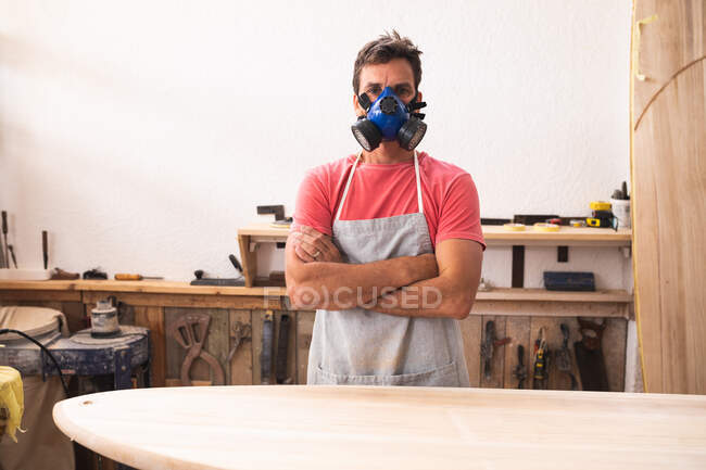 Porträt eines kaukasischen Surfbrettmachers, der in seinem Atelier arbeitet, eine Schutzschürze und eine Gesichtsmaske trägt, mit verschränkten Armen dasteht und in die Kamera blickt. — Stockfoto