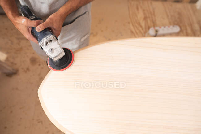 Parte centrale della tavola da surf maschile che lavora nel suo studio, indossando un grembiule protettivo, modellando una tavola da surf in legno con una levigatrice. — Foto stock