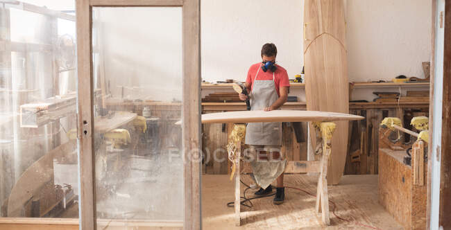 Creatore caucasico di tavole da surf maschile che lavora nel suo studio, indossando un grembiule protettivo e una maschera respiratoria, modellando una tavola da surf in legno con una levigatrice. — Foto stock