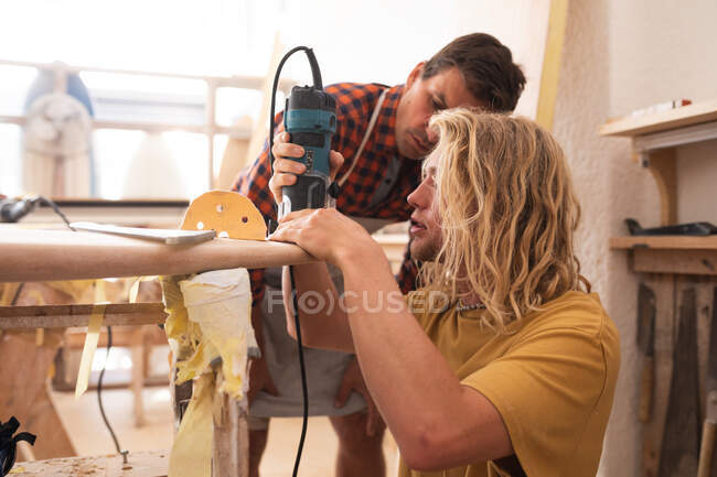 Due surfisti maschi caucasici che lavorano nel loro studio e costruiscono insieme una tavola da surf in legno, lucidandola e modellandola con una levigatrice. — Foto stock