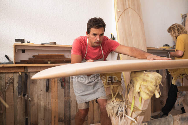 Dos surfistas caucásicos trabajando en su estudio y haciendo una tabla de surf de madera juntos, inspeccionándola antes de darle forma con una lijadora. - foto de stock