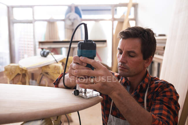 Fabricante de tablas de surf masculino caucásico trabajando en su estudio, usando un delantal protector, formando una tabla de surf de madera con una lijadora. - foto de stock