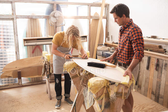 Dos surfistas caucásicos trabajando en su estudio y haciendo una tabla de surf de madera juntos, puliendo y limpiando la superficie. - foto de stock