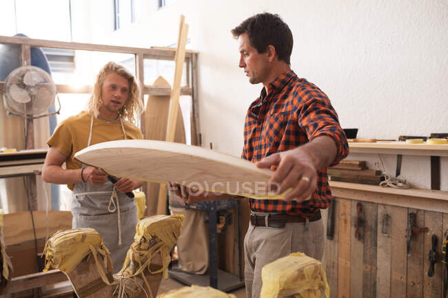 Zwei kaukasische männliche Surfbrettmacher arbeiten in ihrem Atelier und stellen gemeinsam ein hölzernes Surfbrett her, inspizieren es, bevor sie die Oberfläche lackieren. — Stockfoto