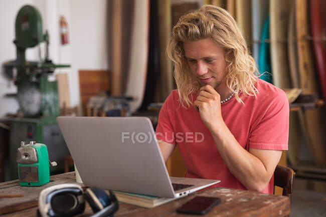 Produttore caucasico di tavole da surf maschile che lavora nel suo studio, seduto alla scrivania e utilizzando il suo computer portatile, con tavole da surf in un rack sullo sfondo — Foto stock