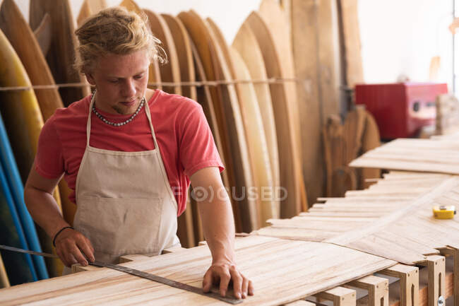 Creatore di tavole da surf caucasiche che lavora nel suo studio, taglia strisce di legno e si prepara a fare una tavola da surf, con tavole da surf in un rack sullo sfondo — Foto stock