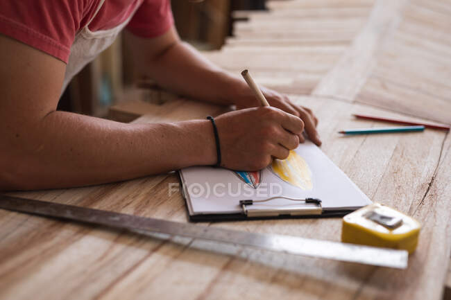 Sección media del fabricante de tablas de surf masculino trabajando en su estudio, dibujando proyectos de tablas de surf en un cuaderno de bocetos, preparándose para hacer una tabla de surf. - foto de stock