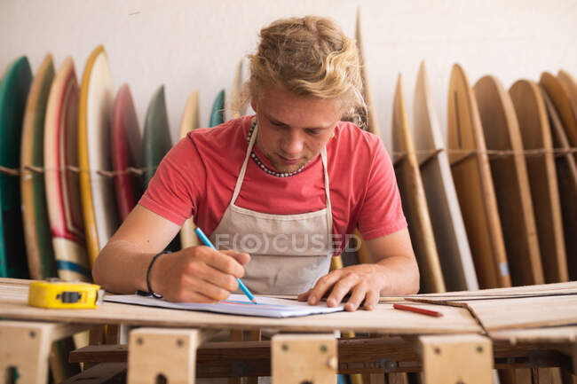 Homme blanc fabricant de planches de surf travaillant dans son studio, dessinant des projets de planches de surf dans un carnet de croquis, avec planches de surf dans un rack en arrière-plan. — Photo de stock