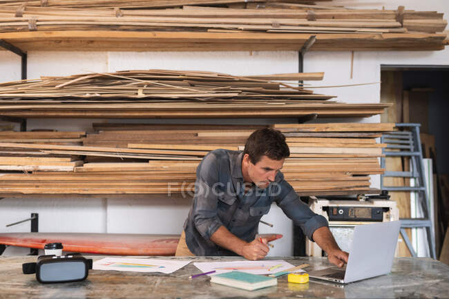 Caucasian male surfboard maker working in his studio, standing behind the counter and using his laptop computer while preparing a surfboard project, with wooden strips hanging in the background. — Stock Photo