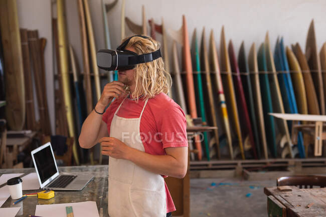 Caucasian male surfboard maker working in his studio, using a VR headset, with surfboards in a rack in the background. — Stock Photo