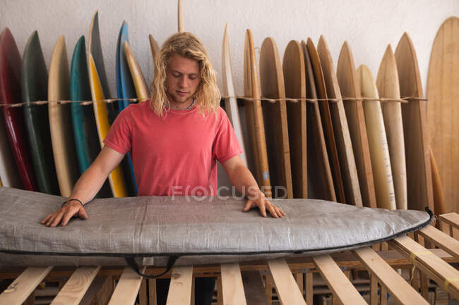 Fabricante de tablas de surf masculino caucásico trabajando en su estudio, inspeccionando una tabla de surf cubierta con una caja gris, con tablas de surf en un estante en el fondo. - foto de stock