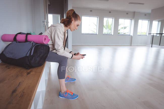 Caucasian attractive female ballet dancer with red hair wearing sportswear, sitting on a bench, preparing for a ballet class, looking at her phone with earphones in. — Stock Photo