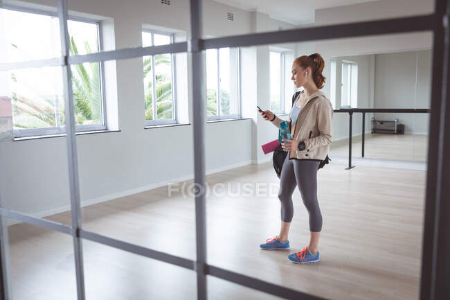 Caucasian attractive female ballet dancer with red hair wearing sportswear, entering a studio, preparing for a ballet class, looking at her phone with earphones in. — Stock Photo