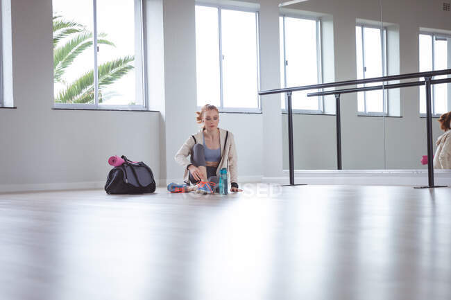 Кавказская привлекательная балетная танцовщица с рыжими волосами в спортивной одежде, входит в студию, готовится к балетному классу, завязывает шнурки, сидит на полу. — стоковое фото
