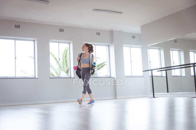 Ballerina caucasica attraente con i capelli rossi che indossa abbigliamento sportivo, entra in uno studio, si prepara per una lezione di balletto, tiene una bottiglia con acqua. — Foto stock