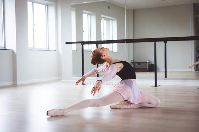 Caucasian attractive female ballet dancer with red hair stretching out, preparing for a ballet class in a bright studio, focusing on her exercise, sitting on the floor. — Stock Photo