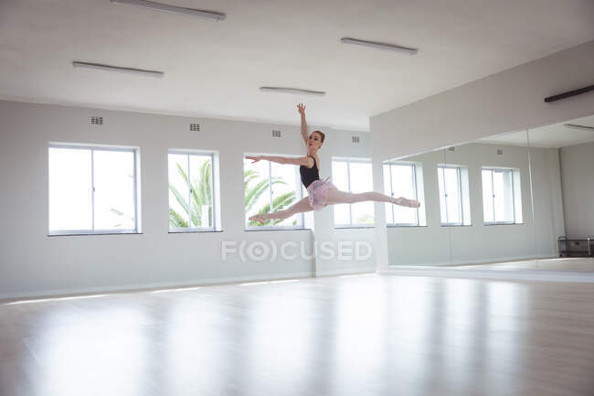 Caucasian attractive female ballet dancer with red hair dancing ballet, preparing for a ballet class in a bright studio, focusing on her exercise jumping in the air with her arm above her head. — Stock Photo