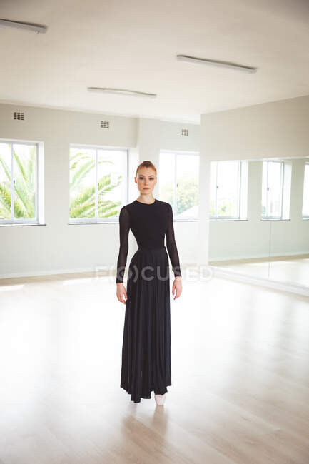 Portrait of an attractive Caucasian female ballet dancer with red hair in a bun wearing a long, black dress, standing on her toes in pointe shoes in a bright ballet studio, looking straight to camera — Stock Photo
