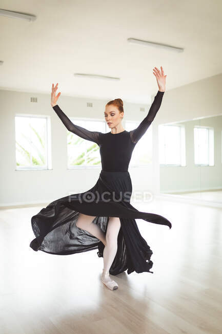 Привлекательная кавказская танцовщица балета с рыжими волосами, танцующая в длинном, черном платье, практикующая балет в яркой студии, фокусирующаяся на своих упражнениях. — стоковое фото