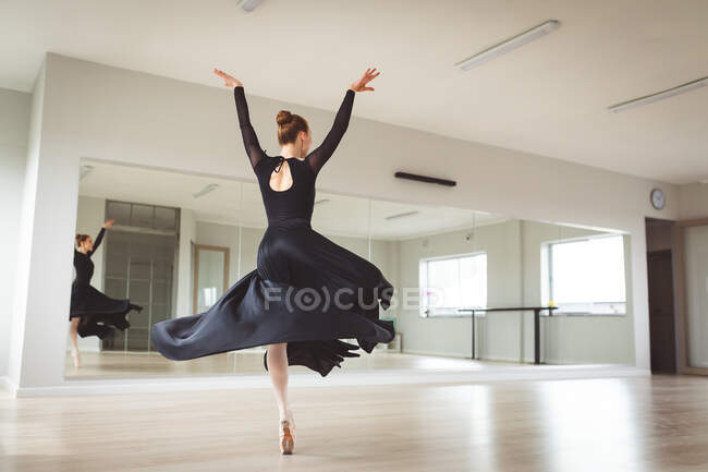 Caucasiana atraente dançarina de balé feminino com cabelo vermelho dançando balé, vestindo um vestido preto, longo, se preparando para uma aula de balé em um estúdio brilhante, com foco em seu exercício . — Fotografia de Stock
