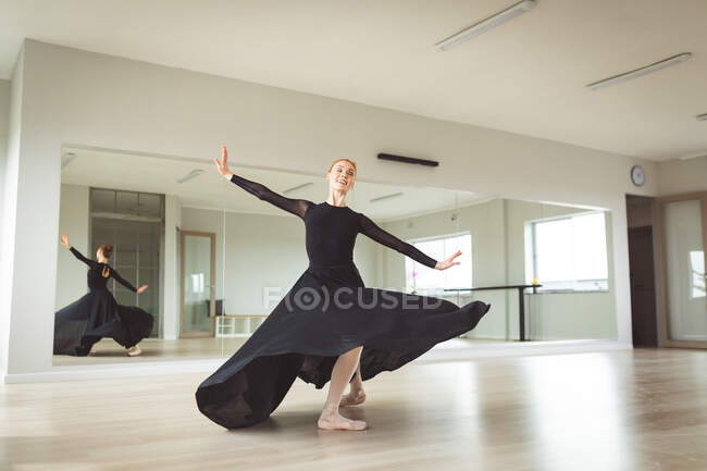 Кавказская привлекательная балетная танцовщица с рыжими волосами танцует балет, одета в черное, длинное платье, готовится к балетному классу в яркой студии, фокусируется на своих упражнениях, улыбается. — стоковое фото
