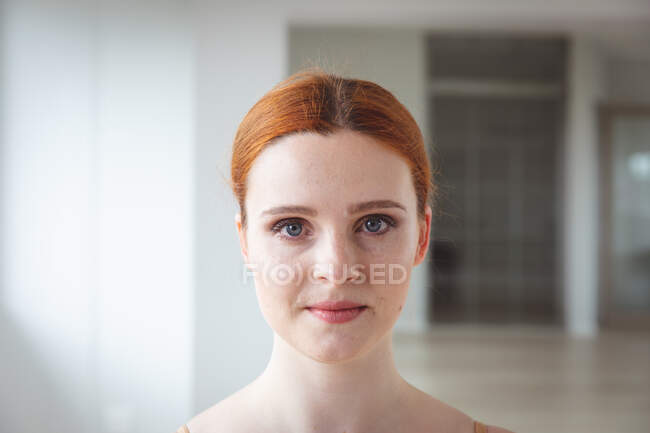 Retrato de una atractiva bailarina de ballet caucásica con el pelo rojo preparándose para una clase de ballet en un estudio brillante, mirando a la cámara con una sonrisa en la cara. - foto de stock