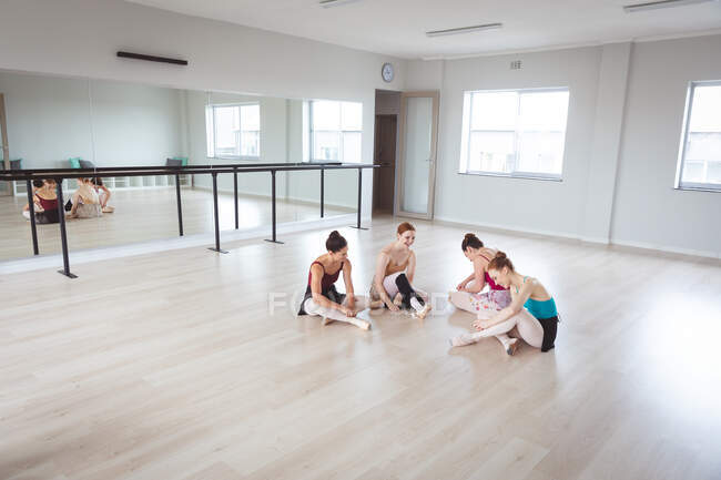 Eine Gruppe kaukasischer attraktiver Balletttänzerinnen unterhält sich in einem hellen Ballettstudio, sieht glücklich aus, bereitet sich auf einen Ballettkurs vor, dehnt sich und sitzt auf dem Boden. — Stockfoto