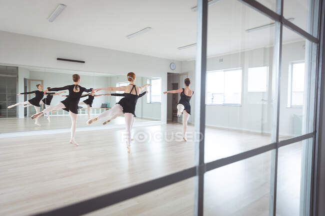 Um grupo de dançarinas de balé caucasianas em roupas pretas praticando durante uma aula de balé dançando na frente de um espelho em um estúdio brilhante, visto através de uma janela . — Fotografia de Stock