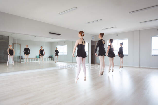 Eine Gruppe attraktiver kaukasischer Balletttänzerinnen in schwarzen Anzügen übt während eines Ballettkurses in einem hellen Studio, tanzt vor einem Spiegel und springt im Gleichklang — Stockfoto
