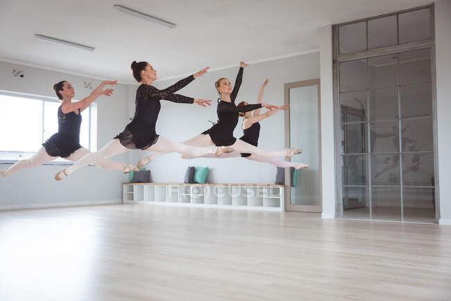 Un grupo de bailarinas caucásicas atractivas en trajes negros practicando durante una clase de ballet en un estudio brillante, bailando y saltando al aire al unísono. - foto de stock