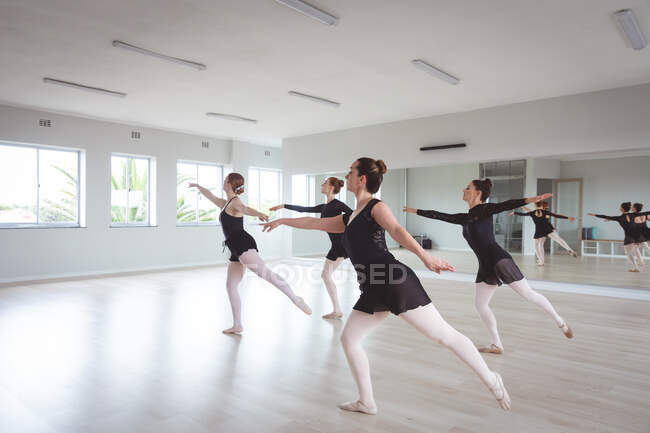 Група кавказьких танцюристок в чорному вбранні під час класу балету в яскравій студії танцюють в унісон перед дзеркалом.. — стокове фото
