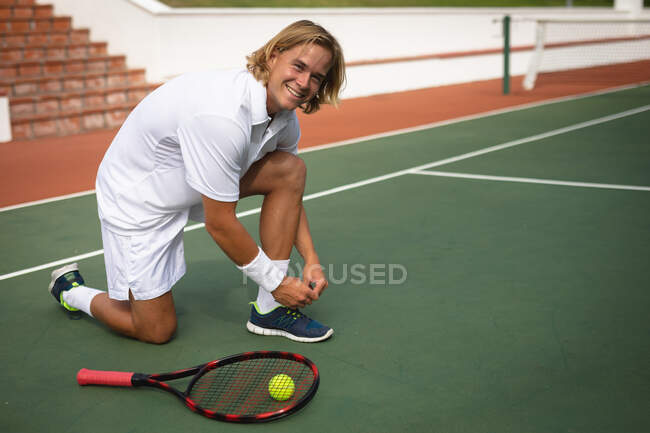 Retrato de un hombre caucásico vistiendo blancos de tenis pasando tiempo en una cancha jugando al tenis en un día soleado, atando cordones de zapatos, mirando a la cámara y sonriendo - foto de stock