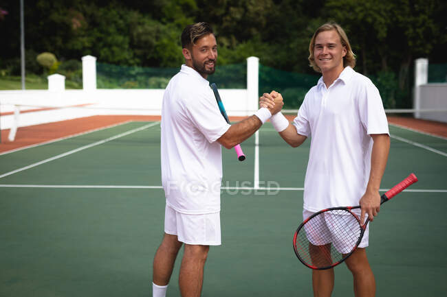 Retrato de um caucasiano e um mestiço vestindo brancos de tênis passando tempo em um tribunal juntos, jogando tênis em um dia ensolarado, apertando as mãos, olhando para a câmera e sorrindo — Fotografia de Stock