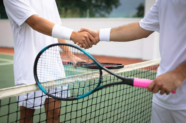 Sección media primer plano de los hombres que usan blancos de tenis pasar tiempo en una cancha juntos, jugar al tenis en un día soleado, estrechando la mano, sosteniendo una raquetas de tenis - foto de stock