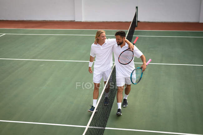 A Caucasian and a mixed race men wearing tennis whites spending time on a court together, playing tennis on a sunny day, embracing and smiling, holding tennis rackets — Stock Photo