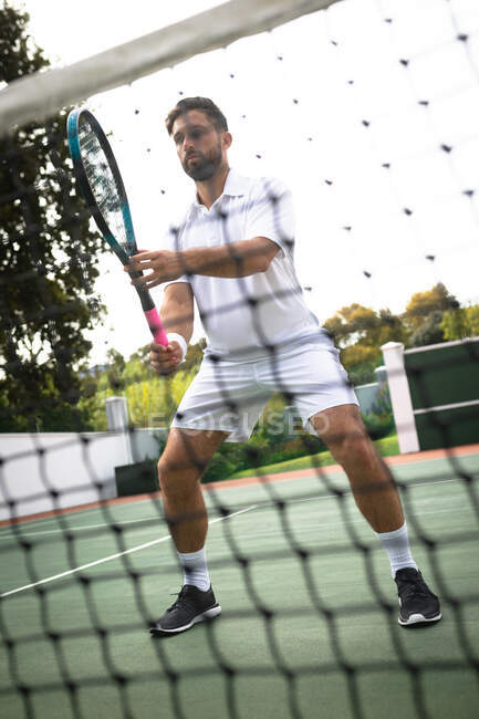 Un hombre de raza mixta vistiendo blancos de tenis que pasa tiempo en una cancha jugando al tenis en un día soleado, sosteniendo una raqueta de tenis, con una red en primer plano - foto de stock