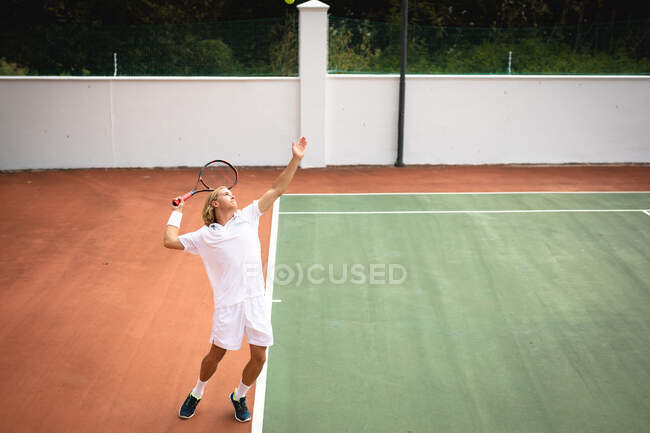 Un hombre caucásico vistiendo blancos de tenis pasando tiempo en una cancha jugando tenis en un día soleado, sosteniendo una raqueta de tenis y preparándose para golpear una pelota - foto de stock