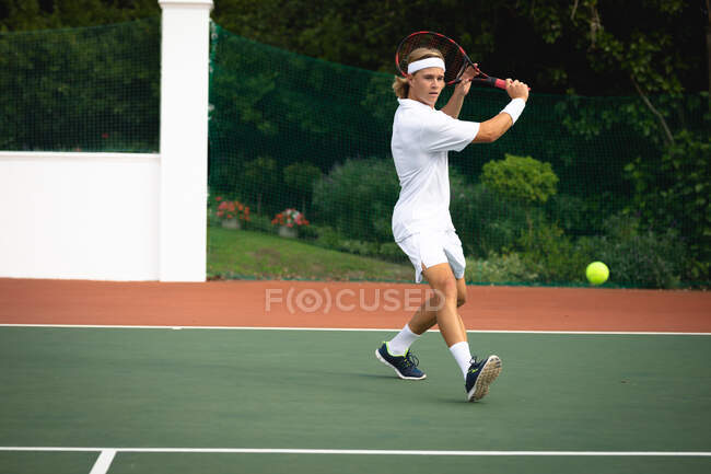Кавказький чоловік у тенісному білому одязі проводить час на корті, граючи в теніс у сонячний день, тримаючи тенісний рекет і готуючись ударити по м 