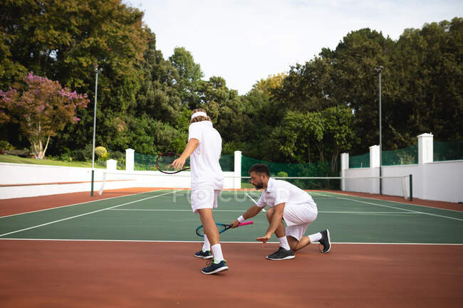 Ein kaukasischer und ein gemischter Rasse Männer in weißen Tennisanzügen verbringen Zeit auf einem Platz zusammen, spielen Tennis an einem sonnigen Tag und halten Tennisschläger in der Hand — Stockfoto