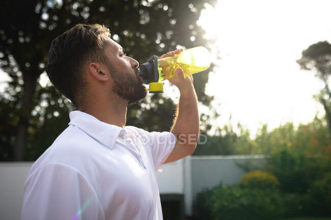 Un hombre de raza mixta con ropa blanca de tenis que pasa tiempo en una cancha jugando al tenis en un día soleado, tomando un descanso y bebiendo agua de una botella. - foto de stock