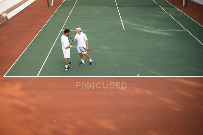 Ein kaukasischer und ein gemischter Rasse Männer in weißen Tennisanzügen verbringen Zeit auf einem Platz zusammen, spielen Tennis an einem sonnigen Tag und halten Tennisschläger in der Hand — Stockfoto