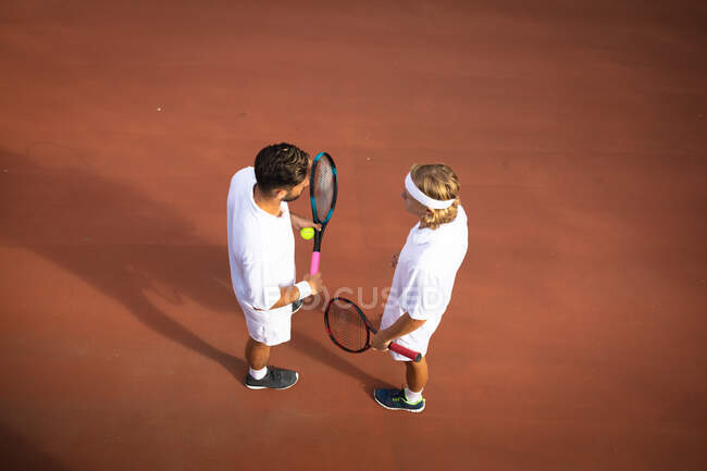 Um caucasiano e um misto vestindo brancos de tênis passando tempo em um campo juntos, jogando tênis em um dia ensolarado, segurando raquetes de tênis e uma bola — Fotografia de Stock