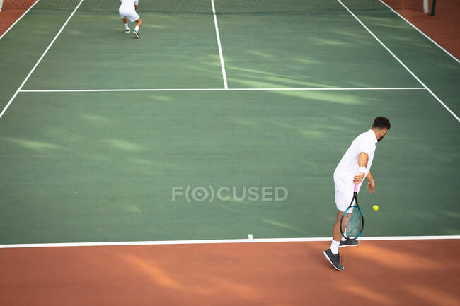 Un caucásico y un hombre de raza mixta con ropa blanca de tenis pasando tiempo en una cancha juntos, jugando tenis en un día soleado, uno de ellos preparándose para golpear una pelota - foto de stock