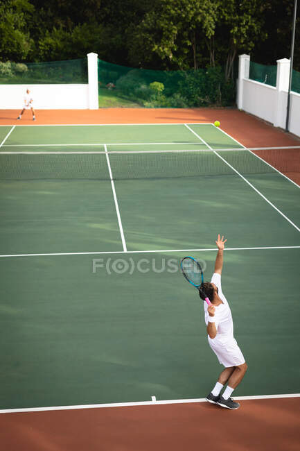 Ein kaukasischer und ein gemischter Rasse Männer in weißen Tennisanzügen verbringen Zeit auf einem Platz zusammen, Tennis spielen an einem sonnigen Tag, einer von ihnen bereitet sich auf einen Ball — Stockfoto