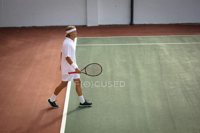 Um homem caucasiano usando tênis branco passando tempo em um campo de ténis em um dia ensolarado, segurando uma raquete de tênis — Fotografia de Stock
